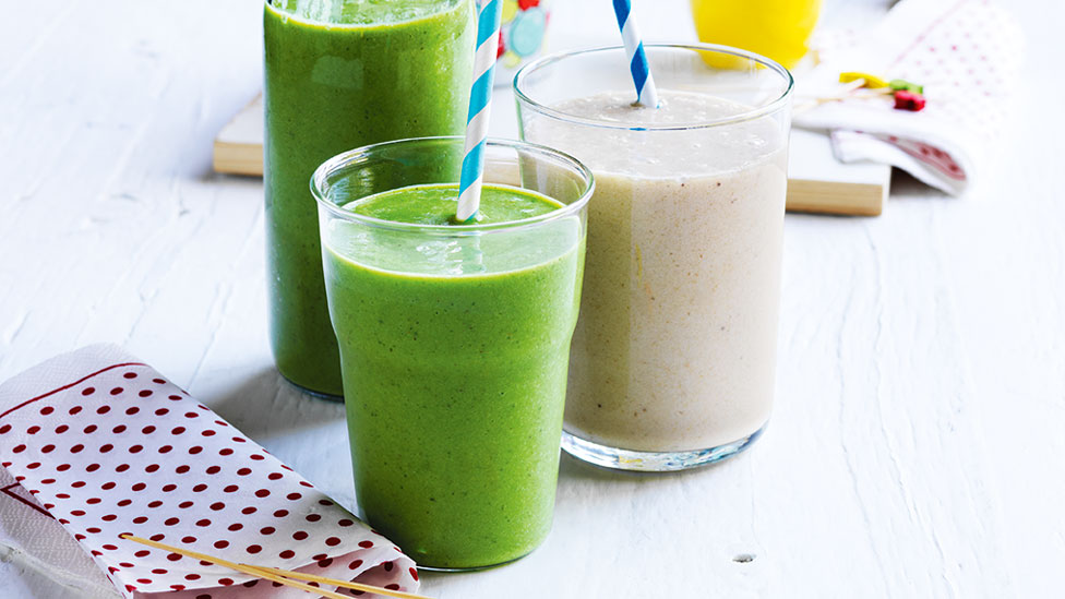 Spinach smoothies in glasses, with a jug of smoothie and chopped fruit on side